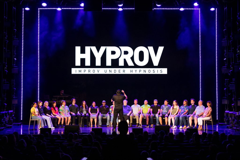 Performer on stage, using hypnosis to entertain an audience, in front of him are 20-25 people on stage he is hypnotzing. The man is about 6 ft tall with a black t-shirt. He has his back on the camera and is facing the people he is hypnotizing. 