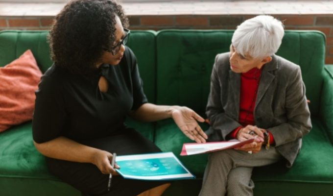 Two women on a green couch talking about pricing and reviewing documents.