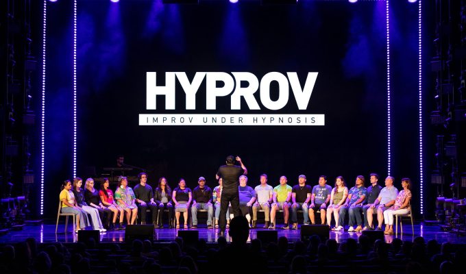 Performer on stage, using hypnosis to entertain an audience, in front of him are 20-25 people on stage he is hypnotzing. The man is about 6 ft tall with a black t-shirt. He has his back on the camera and is facing the people he is hypnotizing.