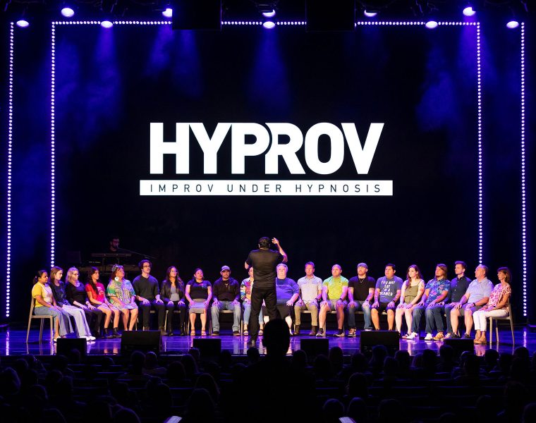 Performer on stage, using hypnosis to entertain an audience, in front of him are 20-25 people on stage he is hypnotzing. The man is about 6 ft tall with a black t-shirt. He has his back on the camera and is facing the people he is hypnotizing.